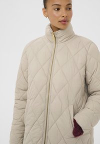 Beige quiltet jakke med høj krave, guld lynlås, sidelommer og burgunder strikdetaljer på manchetterne. Glat, tekstureret stoffinish.