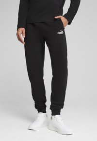 Sorte sweatpants lavet af en blanding af bomuld, med elastisk talje, tapered ben og et hvidt Puma-logo på venstre lår. Sammen med hvide sneakers.