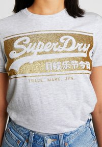 Šedé bavlněné tričko s grafikou ve zlatém třpytu s nápisem "SuperDry" a japonským textem pod ním. Krátké rukávy a kulatý výstřih.