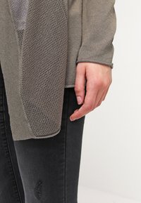 Grå cardigan med en texturerad, stickad design och en slät ärm. Buren över svarta jeans, vilket visar en detaljerad hand och handled.