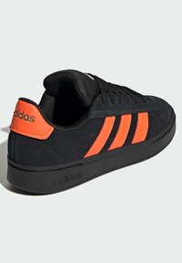 Sneaker nera Adidas con linguetta del tallone arancione brillante e tre strisce laterali, lacci neri e suola marchiata su uno sfondo chiaro.