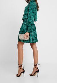 Robe en imprimé léopard teal, taille cintrée, manches longues. Talons hauts noirs à brides. Tenant une pochette argentée étincelante. Texture de tissu lisse.