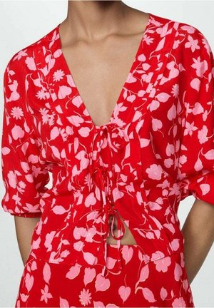 Blouse rouge à imprimé floral rose, encolure en V, manches courtes bouffantes et liens à l'avant, portée avec un bas assorti à motif floral.