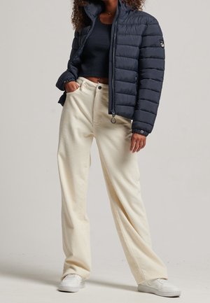 Persoon draagt een marineblauw doorgestikt jack over een zwart cropped top met offwhite wijde broekspijpen en witte sneakers, staat met één hand in de zak.