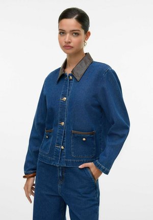 Jeansjacke in Dunkelblau mit einem Kragen aus braunem Kunstleder, goldenen Knöpfen und zwei vorderen Taschen mit braunen Akzenten.