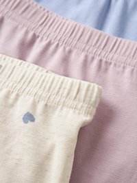 Gestapelde zachte stoffen tailles in crème met een blauw hartdetail en effen lichtpaars, tegen een lichtblauwe achtergrond.