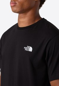 Mand iført sort T-shirt med korte ærmer og hvidt The North Face-logo på venstre bryst, mod en ensfarvet baggrund.