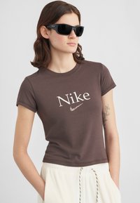 Καφέ μπλουζάκι Nike με λευκό λογότυπο, κοντά μανίκια και κοντή σχεδίαση. Το μοντέλο φοράει μαύρα γυαλιά ηλίου και ανοιχτόχρωμο παντελόνι.