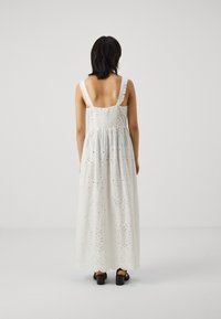 Fehér maxi ruha broderie anglaise mintával, fodros vállpántokkal, rugalmas mellrésszel és réteges szegéllyel. Fekete tömbsarúval viselve.