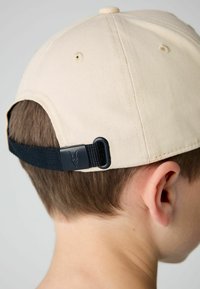 Beige baseballsapka fekete pánttal és csattal a hátsó részen. A tetején gomb és szellőzőnyílások találhatóak a koronán.
