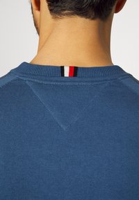 Suéter de malha azul com um tecido texturizado. Gola canelada com riscas vermelha, preta e branca na parte de trás. Inclui um detalhe de costura em forma de V.