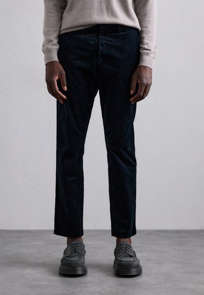 Pantaloni din catifea întunecată, cu o croială slim, având un front plat și o textură subtilă. Asortați cu pantofi gri cu șiret.