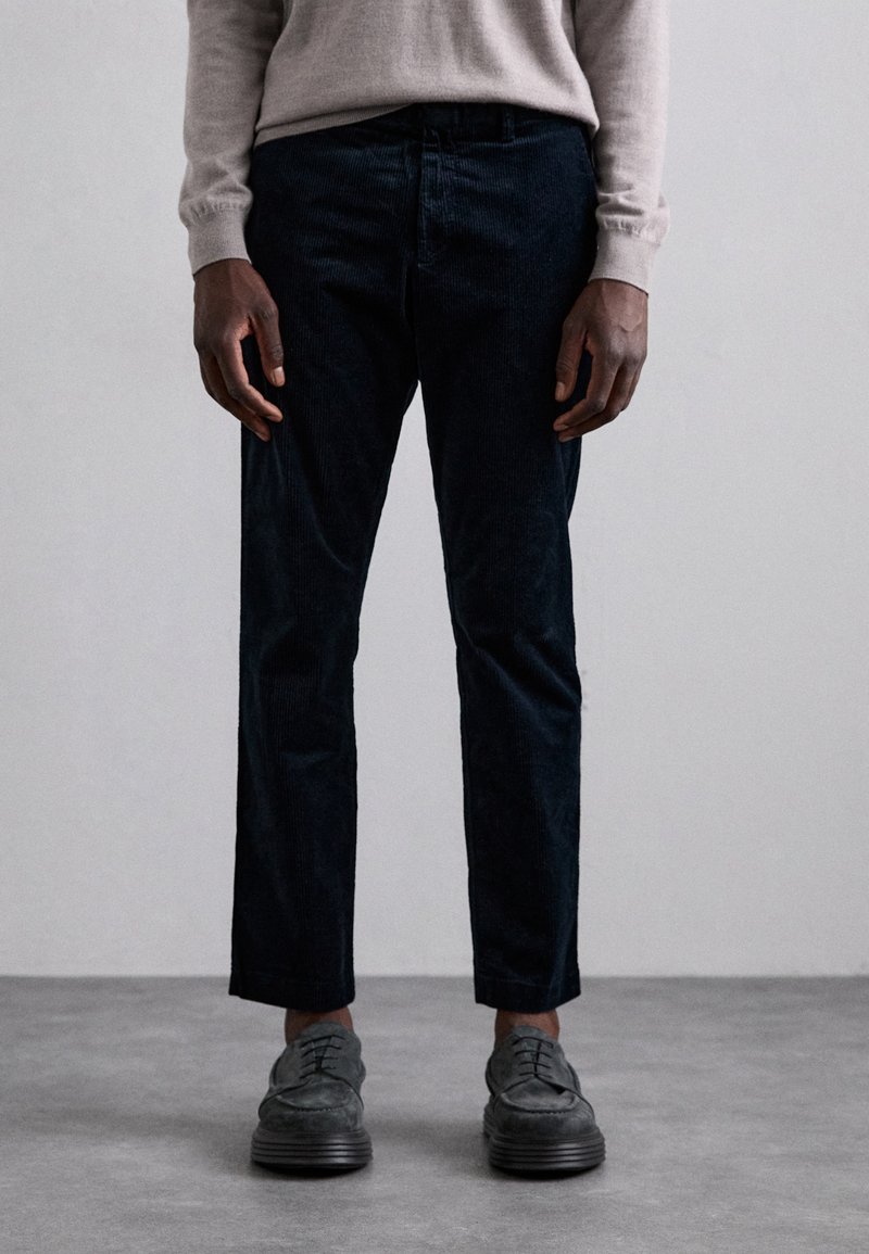 Dark corduroy trousers with a slim fit, featuring a flat front and subtle texture. Paired with gray lace-up shoes.