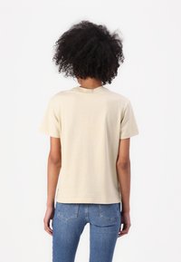 Beige cotton t-shirt with a rounded neckline and short sleeves, featuring a simple design. Paired with blue denim jeans, showing a straight cut.