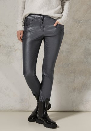 Persona con pantalones ajustados gris oscuro brillante, botines negros de charol y un suéter de punto de color claro, con una mano en el bolsillo.