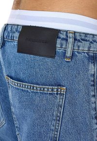 Denimfarkut keskisinisessä pesussa, sileä pinta. Takana musta nahkainen merkki, näkyvät vyölenkit ja kontrastiset ompeleet.