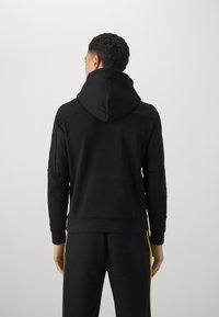 Felpa nera con un grande cappuccio, con pannelli laterali testurizzati e polsini a coste. Abbinata a pantaloni neri coordinati e strisce gialle sui lati.