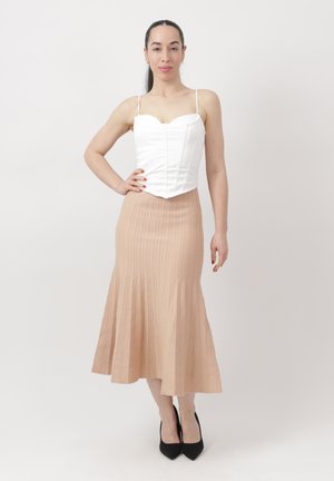 Kobieta z długimi włosami, ubrana w białą bezrękawnik i beżową plisowaną spódnicę, stojąca z jedną ręką na biodrze i nosząca czarne szpilki.