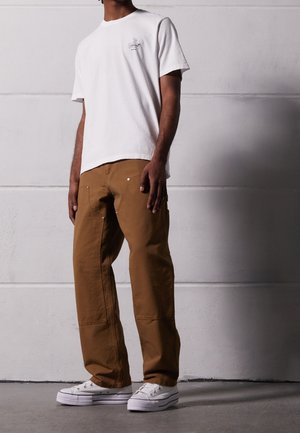 Homme portant un t-shirt blanc à motif, un pantalon de travail marron avec coutures renforcées et des baskets blanches basses, debout contre un mur uni.