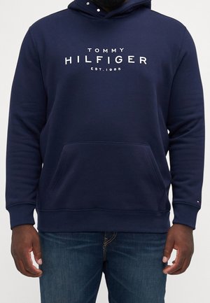 Marineblå hættetrøje med teksten "Tommy Hilfiger Est. 1985", frontlomme, båret af en person med mørk hud og blå jeans.