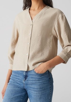 Frau trägt beige langärmelige Bluse mit Knöpfen und blaue Jeans, eine Hand in der Tasche, gezeigt vom Hals bis zur Hüfte vor einfarbigem Hintergrund.