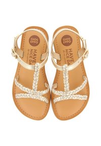 Sandalias de cuero doradas con correas trenzadas y hebilla en el tobillo. Plantilla color tostado con texto de la marca. Diseño de punta abierta, suela plana, estilo minimalista.