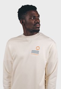 Sudadera de color crema con mangas largas, que presenta un logo frontal de un elefante estilizado y líneas onduladas azules; textura de algodón.