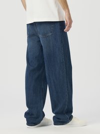 Raspuštene plave traperice od trapera, s opuštenim krojem, dizajnom s pet džepova i bijelim tenisicama, prikazane s leđa.