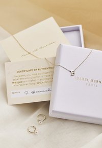 Goldkette mit dem Buchstaben "B", präsentiert in einer weißen Box. Enthält ein Echtheitszertifikat auf cremefarbenem Papier. Goldene Ohranhänger mit Perlen.