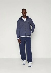 Mornarska zip-up kapuca z belimi črtastimi rokavi in ustreznimi mornarskimi hlačami. Mehak material, sproščen kroj, z izvezenim logotipom na prsih.