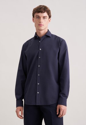 Ung mand iført en mørkeblå skjorte med lange ærmer og knapper, stående foran en ensfarvet lys baggrund og vendt mod fronten.