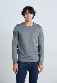 T-shirt gris à manches longues en coton doux avec un col rond. Présente un petit logo rouge sur la poitrine gauche. Assorti à un pantalon bleu marine.