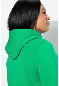 Zelená mikina s kapucí z měkké látky, vybavená stahovací šňůrkou na kapuci, volným střihem a viditelným prošitím podél švů.