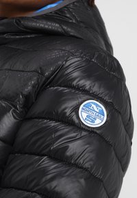 Svart puffjacka med kviltad design, med en rund blå och vit North Sails-logotyp på axeln och en glatt, glänsande yta.