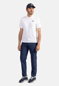 Vit polo med kort ärm, marinblå keps, marinblå byxor och svarta sneakers med vita detaljer och snören, mot en enkel bakgrund.