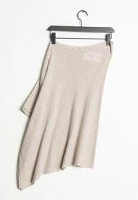 Asymmetrisk beige stickad plagg hängande på en svart galge, med en strukturerad mönstring och en lös, draperad silhuett.