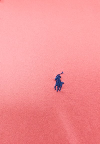 Μπλε κεντημένο λογότυπο παίκτη πόλο πάνω σε άλογο, τοποθετημένο στο κέντρο σε φόντο υφάσματος κοραλλί χρώματος.
