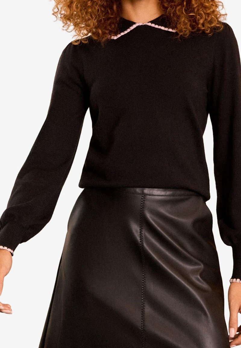 Femme portant un pull noir à manches longues avec une bordure rose et une jupe en cuir noire, posant devant un fond blanc uni.