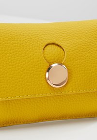 Portefeuille en cuir jaune texturé avec une fermeture métallique ronde dorée sur le rabat avant, posé sur une surface blanche.