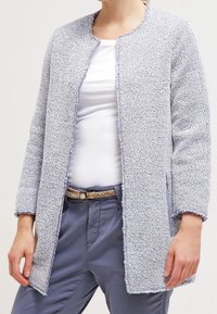 Ljusgrå texturerad cardigan med rund halsringning, öppen framsida och långa ärmar. Bärs över en vit skjorta och kombineras med blå byxor.