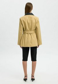 Giacca beige con cintura, colletto nero, maniche lunghe e vestibilità su misura. Abbinata a pantaloni corti neri fino al ginocchio e sandali con tacco.