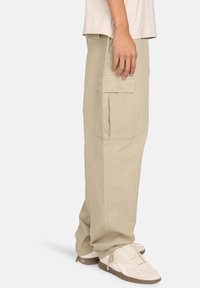 Beige cargobroek met een losse pasvorm, voorzien van grote zijzakken en een gestructureerde stof. Gepaard met beige sneakers en een licht shirt.