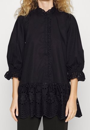 Vrouw draagt een zwarte blouse met opgepofte driekwartsmouwen en kanten oogjes ruches op de manchetten, zoom en voorkant.