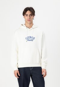 Sudadera blanca con un bolsillo frontal y cordones ajustables. Presenta un logo azul "GUESS JEANS" en un diseño curvado. Combinada con jeans oscuros.