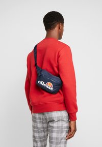 Borsa a spalla blu navy con zip, con logo bianco e dettagli arancioni. Indossata a tracolla sopra un felpa rossa e pantaloni scozzesi.