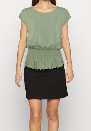 Žena v krátkém rukávu s vrškem šalvějově zelené barvy se staženým pasem a černou sukní délky po kolena na jednobarevném pozadí.