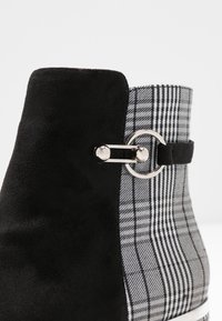 Černé semišové kotníkové boty s kostkovanou látkovou panelem, s kovovým akcentem v podobě stříbrného kroužku na boku. Texturovaný a moderní design.