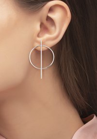 Les boucles d'oreilles en argent présentent un design circulaire avec une tige droite qui intersecte le cercle. Texture lisse, forme minimiste.