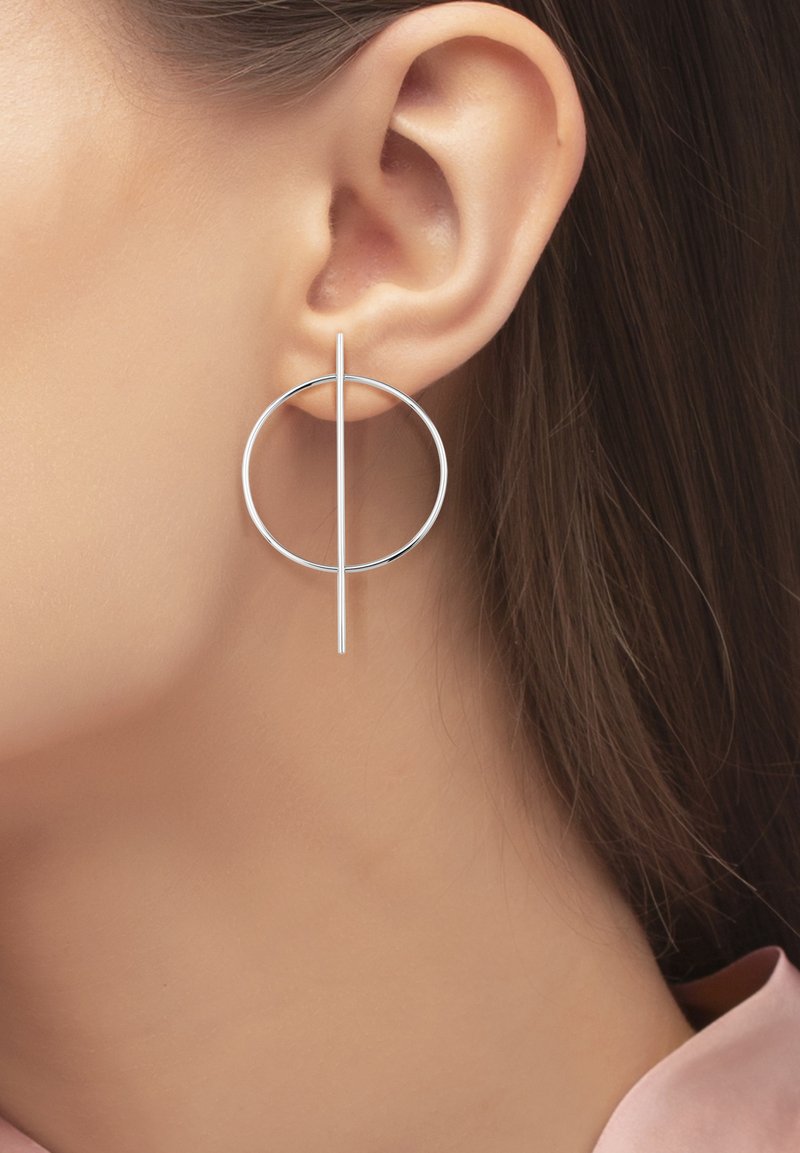 Les boucles d'oreilles en argent présentent un design circulaire avec une tige droite qui intersecte le cercle. Texture lisse, forme minimiste.