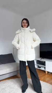 Kurtka puchowa w kolorze kremowym z wysokim kołnierzem, zamkiem błyskawicznym i zatrzaskami, ukośnymi przeszyciami, dużymi kieszeniami i luźnym krojem.